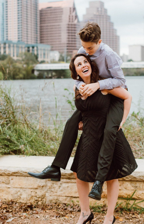 A woman in a black dress gives a piggyback ride to a young person wearing a checkered shirt and dark pants, near a body of water with city buildings in the background.